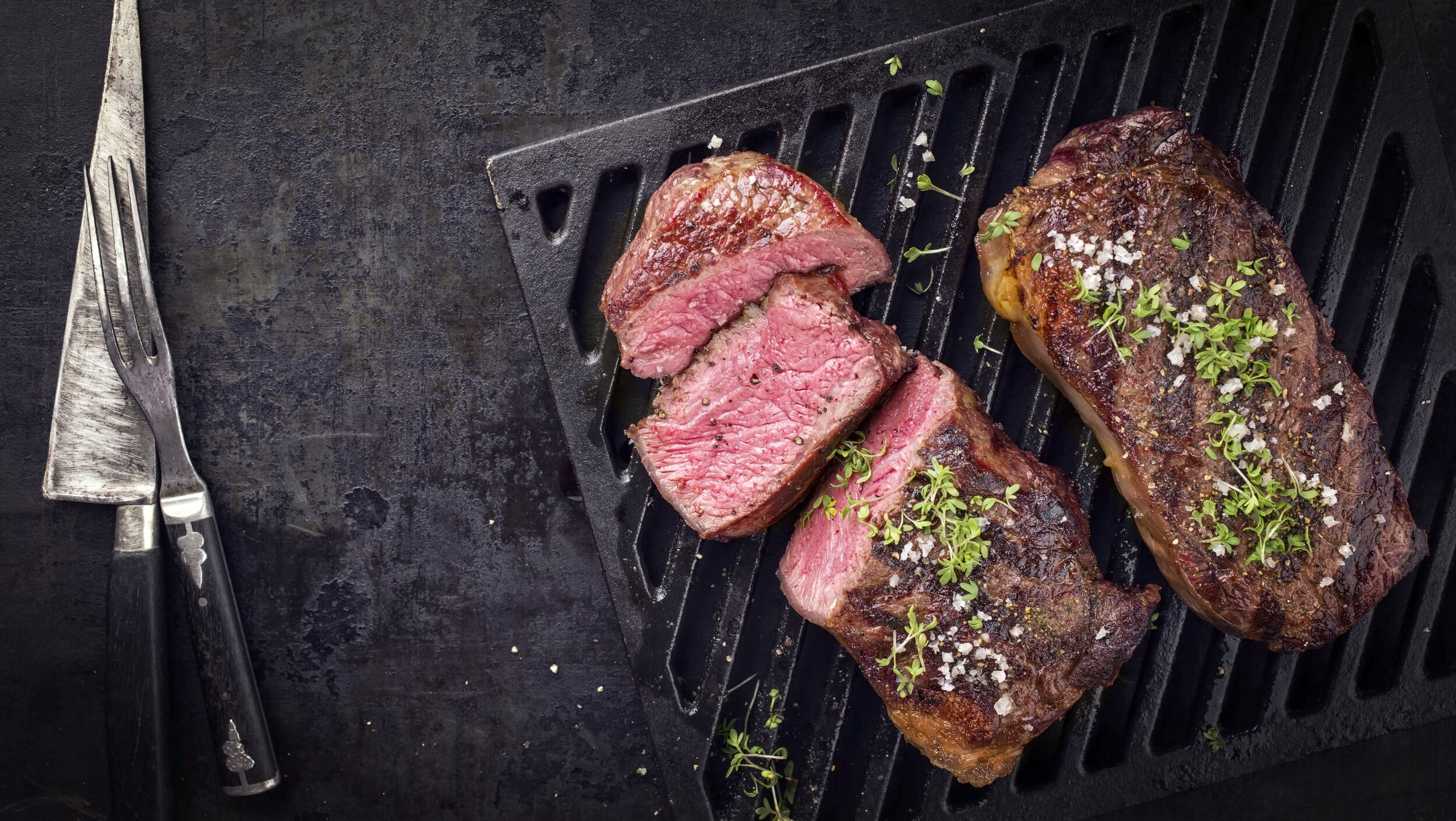 Two barbecue dry aged Kobe roast beef as close-up on a grillage Grilled steaks on a black grilling pan, garnished with fresh herbs and sea salt, accompanied by a silver knife and fork, showcasing juicy pink interiors and a perfectly seared crust.