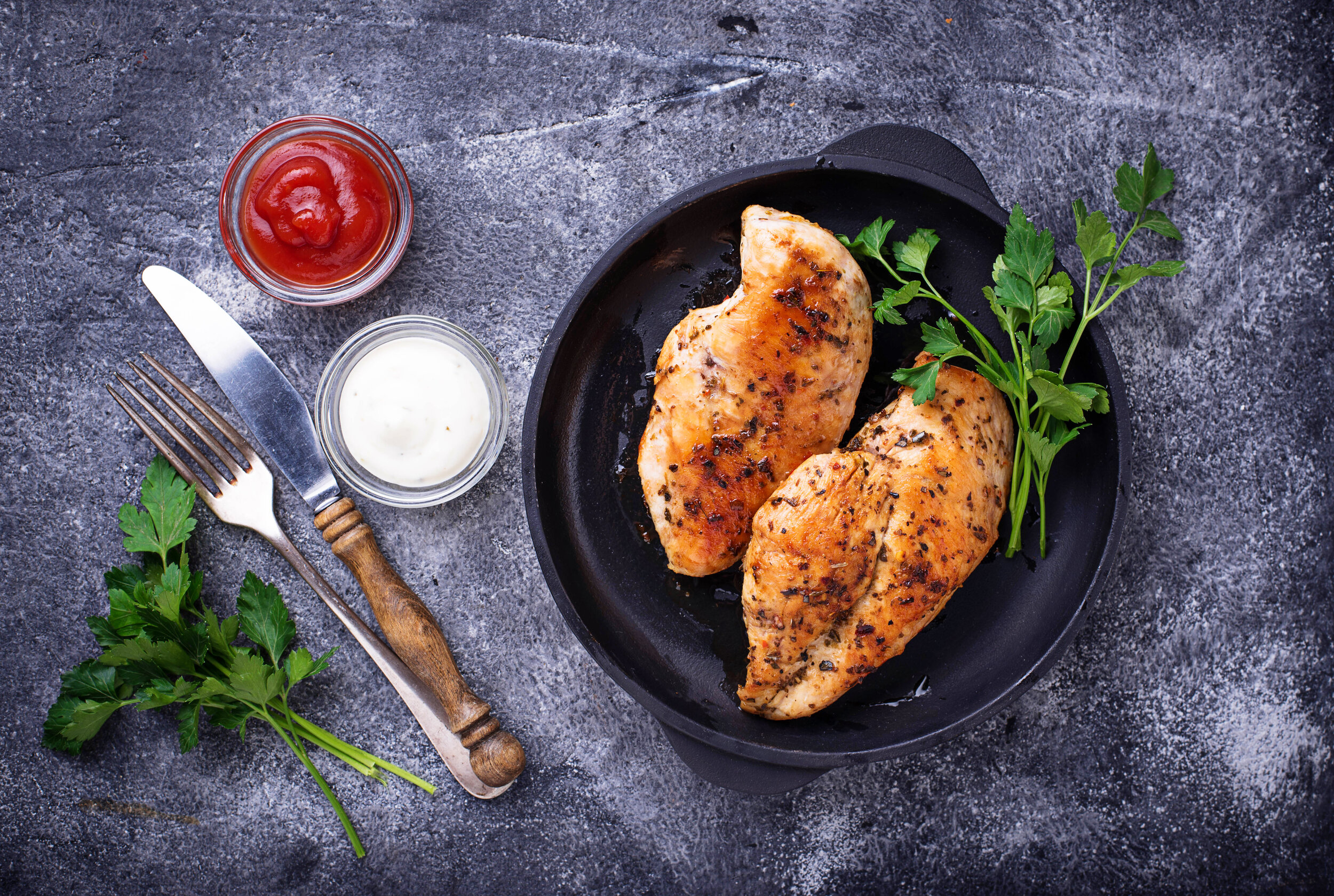 Grilled chicken breast or fillet on iron pan Grilled chicken breasts served in a cast iron skillet, accompanied by fresh parsley and dipping sauces of ketchup and mayonnaise on a dark textured surface.