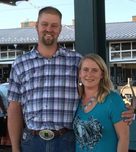 Daniel+West+-+IMG_0156 (1) A man and a woman pose together outdoors, smiling. The man is wearing a plaid shirt and jeans, while the woman is dressed in a teal top with a necklace. They appear to be at a market or outdoor event, with buildings in the background.