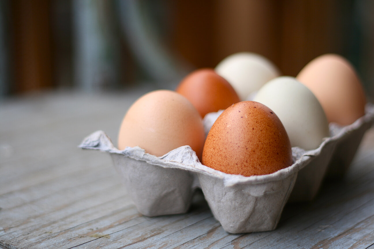 Egg+Carton+7 Carton of assorted eggs in various colors, including brown, white, and tan, placed on a rustic wooden surface. Ideal for culinary and nutrition topics.